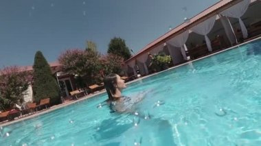 Siyah mayo giymiş genç bir kadın ve açık havada yüzme havuzunda mavi fayanslarla süslenmiş beyaz bir pelerin yüzüyor..