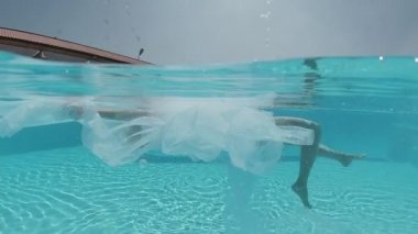 Siyah mayo giymiş genç bir kadın ve açık havada yüzme havuzunda mavi fayanslarla süslenmiş beyaz bir pelerin yüzüyor..
