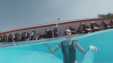 Siyah mayo giymiş genç bir kadın ve açık havada yüzme havuzunda mavi fayanslarla süslenmiş beyaz bir pelerin yüzüyor..