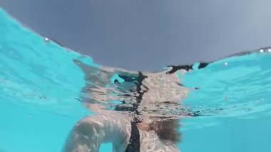 Siyah mayo giymiş genç bir kadın ve açık havada yüzme havuzunda mavi fayanslarla süslenmiş beyaz bir pelerin yüzüyor..
