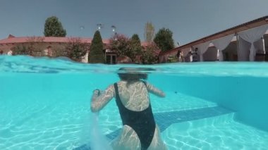 Siyah mayo giymiş genç bir kadın ve açık havada yüzme havuzunda mavi fayanslarla süslenmiş beyaz bir pelerin yüzüyor..