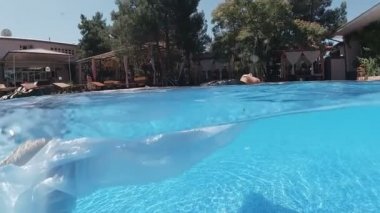 Siyah mayo giymiş genç bir kadın ve açık havada yüzme havuzunda mavi fayanslarla süslenmiş beyaz bir pelerin yüzüyor..