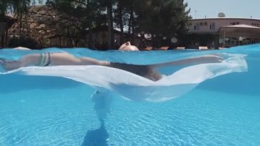 Siyah mayo giymiş genç bir kadın ve açık havada yüzme havuzunda mavi fayanslarla süslenmiş beyaz bir pelerin yüzüyor..