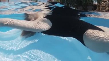 Siyah mayo giymiş genç bir kadın ve açık havada yüzme havuzunda mavi fayanslarla süslenmiş beyaz bir pelerin yüzüyor..