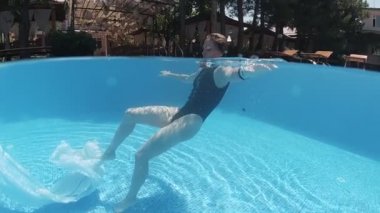 Siyah mayo giymiş genç bir kadın ve açık havada yüzme havuzunda mavi fayanslarla süslenmiş beyaz bir pelerin yüzüyor..
