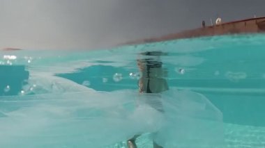 Siyah mayo giymiş genç bir kadın ve mavi fayanslarla süslenmiş havuzun tabanında yürüyen beyaz bir pelerin.