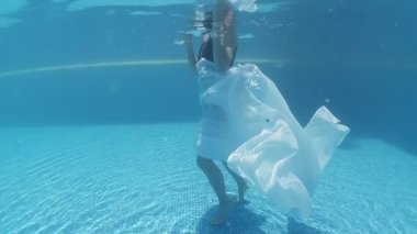 Siyah mayo giymiş genç bir kadın ve mavi fayanslarla süslenmiş havuzun tabanında yürüyen beyaz bir pelerin.