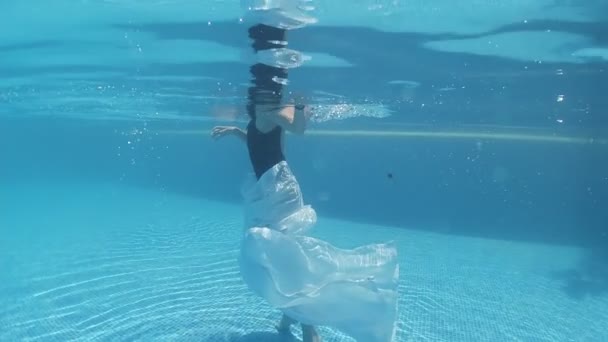 身穿黑色泳衣和白色长袍的年轻女子沿着装饰着蓝色瓷砖的游泳池底部走着 — 图库视频影像