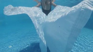 Siyah mayo giymiş genç bir kadın ve mavi fayanslarla süslenmiş havuzun tabanında yürüyen beyaz bir pelerin.