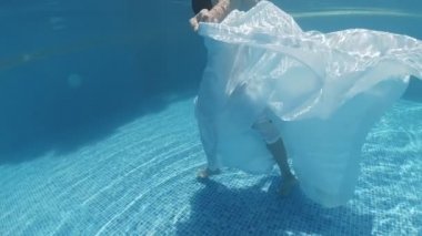 Siyah mayo giymiş genç bir kadın ve mavi fayanslarla süslenmiş havuzun tabanında yürüyen beyaz bir pelerin.