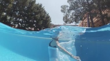 Siyah mayo giymiş genç bir kadın ve mavi fayanslarla süslenmiş havuzun tabanında yürüyen beyaz bir pelerin.