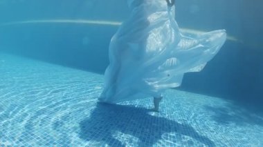 Siyah mayo giymiş genç bir kadın ve mavi fayanslarla süslenmiş havuzun tabanında yürüyen beyaz bir pelerin.