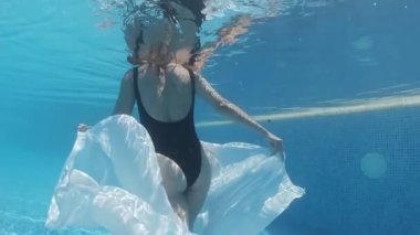 Siyah mayo giymiş genç bir kadın ve mavi fayanslarla süslenmiş havuzun tabanında yürüyen beyaz bir pelerin.