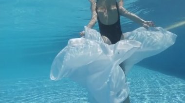 Siyah mayo giymiş genç bir kadın ve mavi fayanslarla süslenmiş havuzun tabanında yürüyen beyaz bir pelerin.