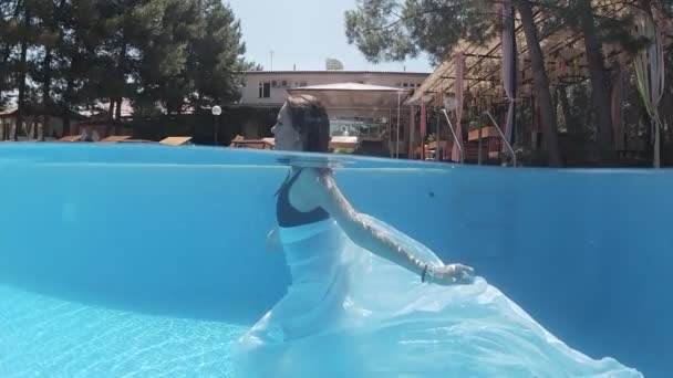 身穿黑色泳衣和白色长袍的年轻女子沿着装饰着蓝色瓷砖的游泳池底部走着 — 图库视频影像