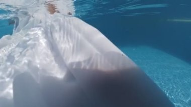Siyah mayo giymiş genç bir kadın ve mavi fayanslarla süslenmiş havuzun tabanında yürüyen beyaz bir pelerin.