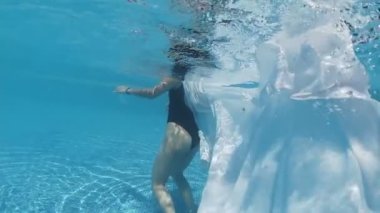 Siyah mayo giymiş genç bir kadın ve mavi fayanslarla süslenmiş havuzun tabanında yürüyen beyaz bir pelerin.