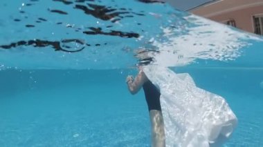 Siyah mayo giymiş genç bir kadın ve mavi fayanslarla süslenmiş havuzun tabanında yürüyen beyaz bir pelerin.