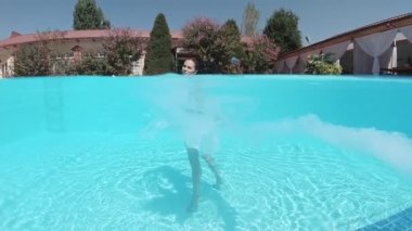 Siyah mayo giymiş genç bir kadın ve mavi fayanslarla süslenmiş havuzun tabanında yürüyen beyaz bir pelerin.