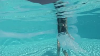 Siyah mayo giymiş genç bir kadın ve mavi fayanslarla süslenmiş havuzun tabanında yürüyen beyaz bir pelerin.