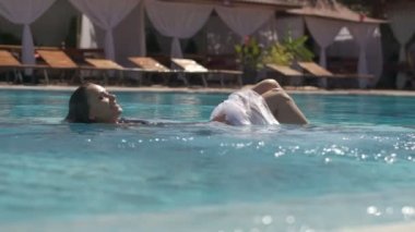 Açık havuzda siyah mayo ve şeffaf pelerin giyen genç bir kadın, ağır çekimde.