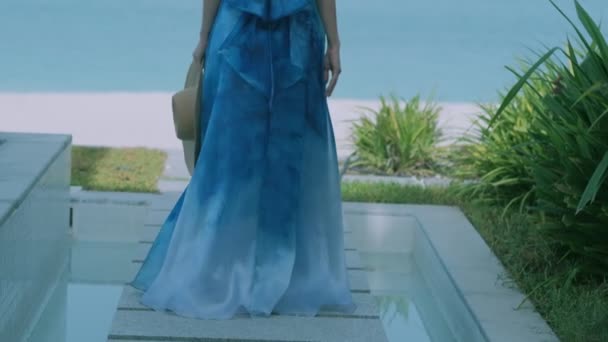 ブロンドの女の子は長い青い夏のドレスを着て海岸線近くの熱帯植物とテラスを歩く スローモーション — ストック動画