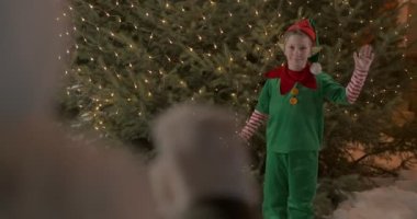 Noel elfi gibi giyinmiş çocuk Noel ağacının önünde el sallıyor, ağır çekimde.