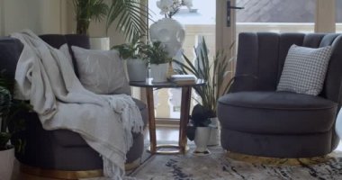 Corner of leaving room with two dark violet armchairs and round coffee table on which plants in pots and books are placed. There are a lot of evergreen plants in the pots in other places. Glass door