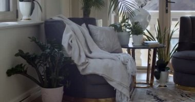 Corner of leaving room with two dark violet armchairs and round coffee table on which plants in pots and books are placed. There are a lot of evergreen plants in the pots in other places. Glass door