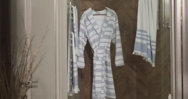 Bathroom tiled with marble tiles in different shades of brown. The plumbing is gold-colored. Baskets with towels is on the floor near big white bathtub. On the left in the frame is a closed toilet
