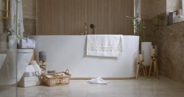 Bathroom tiled with marble tiles in different shades of brown. The plumbing is gold-colored. Baskets with towels is on the floor near big white bathtub. On the left in the frame is a closed toilet