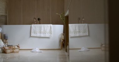 Bathroom tiled with marble tiles in different shades of brown. The plumbing is gold-colored. Baskets with towels is on the floor near big white bathtub. On the left in the frame is a closed toilet