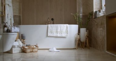 Bathroom tiled with marble tiles in different shades of brown. The plumbing is gold-colored. Baskets with towels is on the floor near big white bathtub. On the left in the frame is a closed toilet