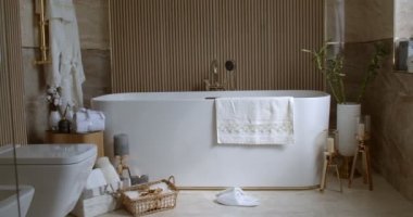 Bathroom tiled with marble tiles in different shades of brown. The plumbing is gold-colored. Baskets with towels is on the floor near big white bathtub. On the left in the frame is a closed toilet