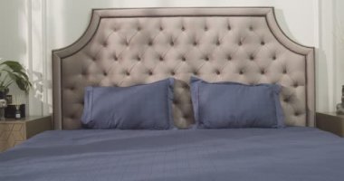 Bedroom filled with soft natural light from the big glass door to the balcony. Large double bed with white linens on the right. Two dark violet armchairs and round coffee table surrounded by evergreen