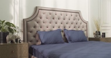 Bedroom filled with soft natural light from the big glass door to the balcony. Large double bed with white linens on the right. Two dark violet armchairs and round coffee table surrounded by evergreen