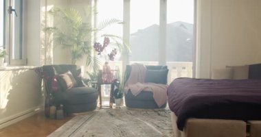 Bedroom filled with soft natural light from the big glass door to the balcony. Large double bed with purple cover on the right. Two armchairs and round coffee table surrounded by evergreen decorative