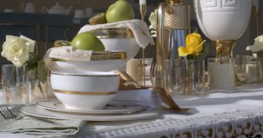 Close-up of dinner table full of dishes and glassware. Dishes are white with gold borders. There are two fruit vases with kiwi and apples. White rose in glass and half of a kiwi are on the left