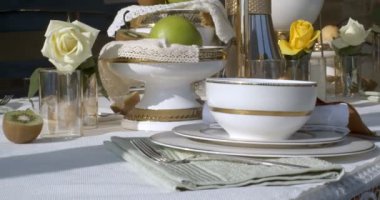 Close-up of dinner table full of dishes and glassware. Dishes are white with gold borders. There are two fruit vases with kiwi and apples. White rose in glass and half of a kiwi are on the left