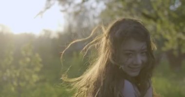Camera follows the girl brown hared girl with grey eyes dressed in airy pink dress walking through apple gardens. Face close-up then close-up of her dress. Filmed against the light from the back.