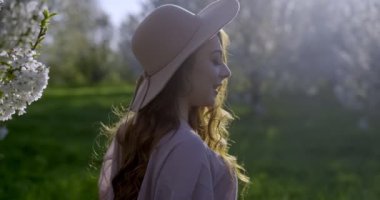 Uzun açık kahverengi saçlı, gri gözlü, pembe elbiseli ve şapkalı bir kız elma bahçelerinde yürüyor. Açık havada çekiliyor, kamera kızı yandan takip ediyor, güneş yüzüne vuruyor..