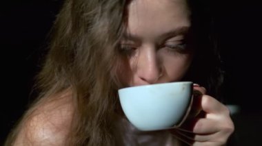 Uzun açık kahverengi saçlı, gri gözlü, beyaz bardaktan zevk alarak kahve içen bir kızın yakın çekimi. Siyah bir arkaplanda tam yüz olarak çekilen