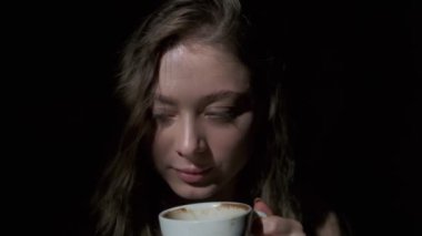 Uzun açık kahverengi saçlı, gri gözlü, beyaz bardaktan zevk alarak kahve içen bir kızın yakın çekimi. Siyah bir arkaplanda tam yüz olarak çekilen
