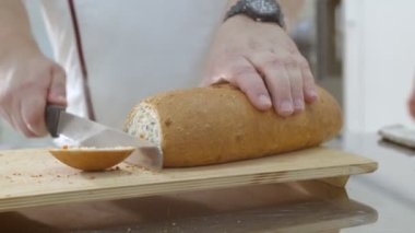 Baker is cutting fresh baked Dutch bread with raisins and dried apricots with bread knife. Crumbs are fell onto the cutting board and table.