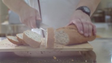 Baker is cutting fresh baked Dutch bread with raisins and dried apricots with bread knife. Crumbs are fell onto the cutting board and table.