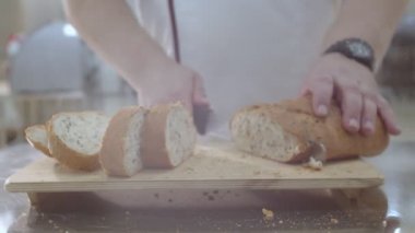 Baker is cutting fresh baked Dutch bread with raisins and dried apricots with bread knife. Crumbs are fell onto the cutting board and table.