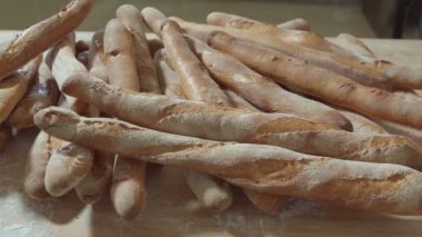 Fresh baked long baguette breads lie on the wooden table sprinkled with flour in a pile. Slow motion