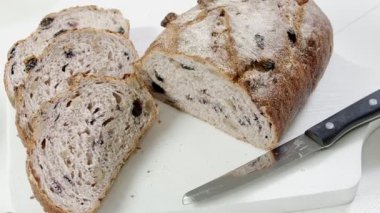 Close-up of sliced rye raisin bread on the white cutting board. There are also fresh veggies, white milk jug and yellow mug on the table. Black handle kitchen knife is on the cutting board. Slightly