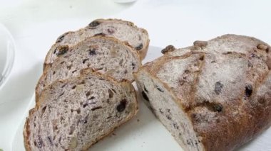 Close-up of sliced rye raisin bread on the white cutting board. There are also fresh veggies, white milk jug and yellow mug on the table. Black handle kitchen knife is on the cutting board. Slightly