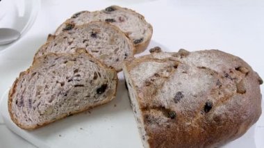 Close-up of sliced rye raisin bread on the white cutting board. There are also fresh veggies, white milk jug and yellow mug on the table. Black handle kitchen knife is on the cutting board. Slightly